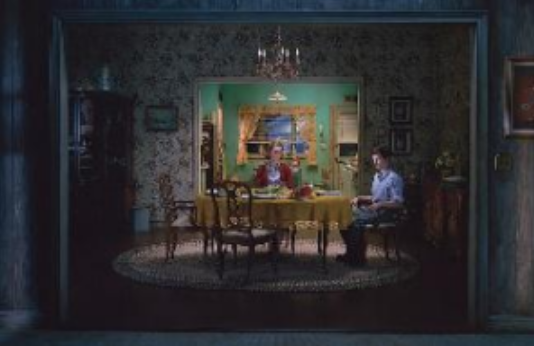 Wohnzimmer mit Esstisch, mehreren Stühlen und zwei sitzenden Personen.